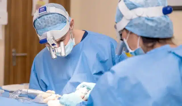 Two surgeons in blue scrubs, masks, and magnifying headgear perform a hair transplant. One operates while the other assists, both focused intently on the delicate procedure. In the background, a door and medical equipment hint at the precision and cost involved in such surgeries.