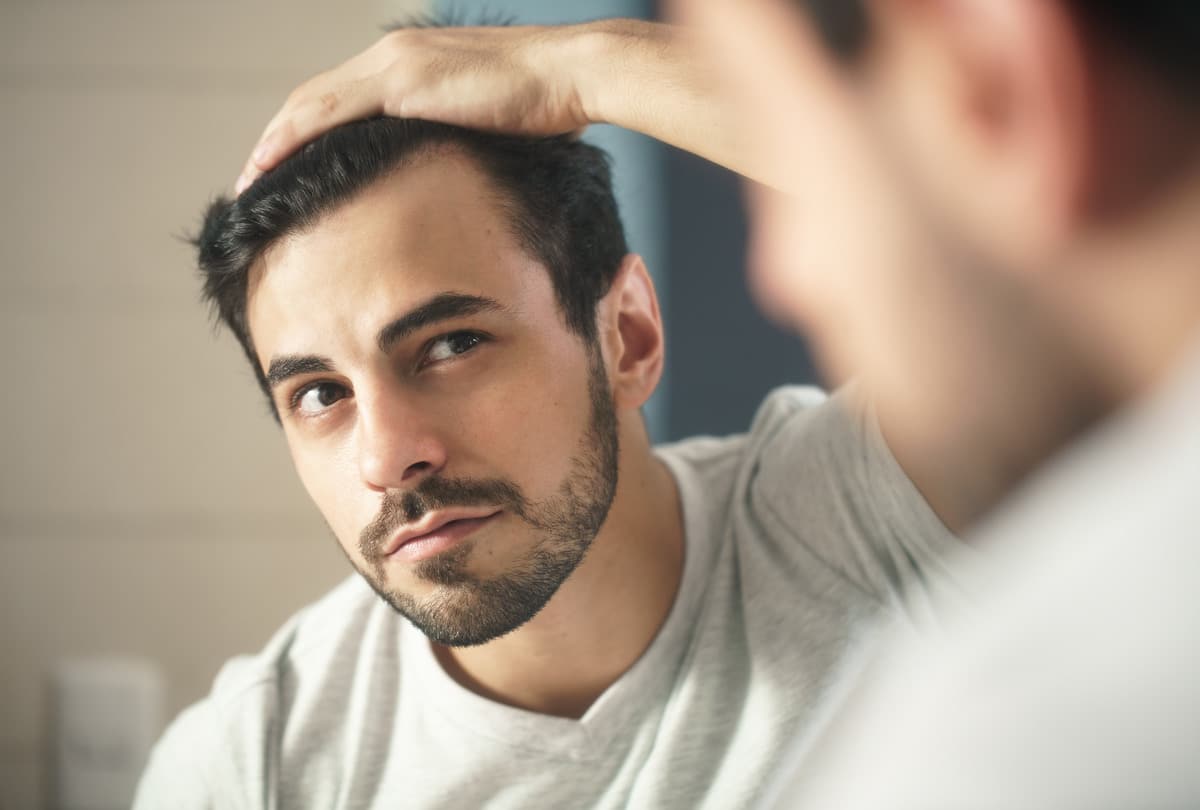 A man with short dark hair and a beard looks in a mirror, examining his hairline. He is wearing a light gray shirt and has an intent expression on his face. The background is softly blurred.