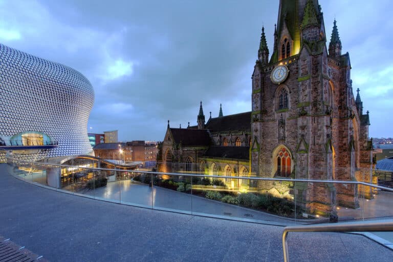 A modern building with a unique, curved pattern, home to a renowned hair loss clinic, stands next to a historic church with a clock tower and Gothic architecture. The scene unfolds at dusk, the sky overcast as city lights begin to illuminate the area.