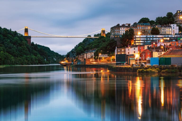 A scenic view at dusk showcases a suspension bridge over the river, with city lights shimmering on the water. Nestled amid lush greenery on the riverbank is a renowned hair loss clinic, all under a cloudy sky's watchful eye.