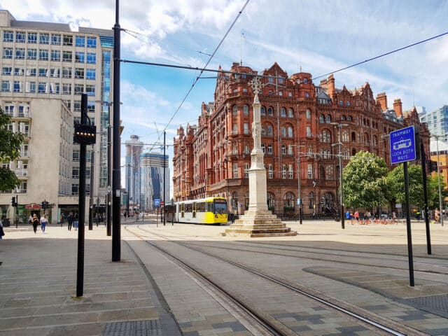 A tram approaches a stop on a cobblestone street in a city plaza. A historic red brick building, now home to a renowned Hair Loss Clinic, stands prominently in the background, alongside modern high rises. Signs and trees line the street under a partly cloudy sky.