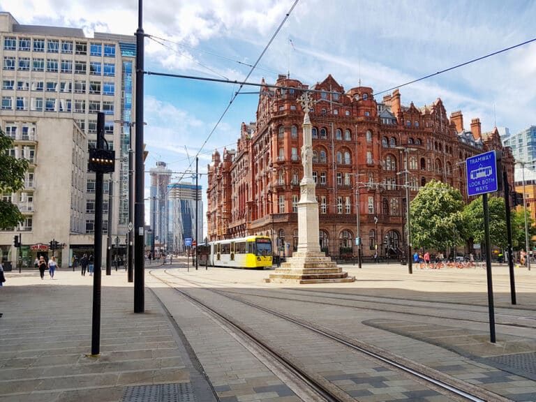 A tram approaches a stop on a cobblestone street in a city plaza. A historic red brick building, now home to a renowned Hair Loss Clinic, stands prominently in the background, alongside modern high rises. Signs and trees line the street under a partly cloudy sky.