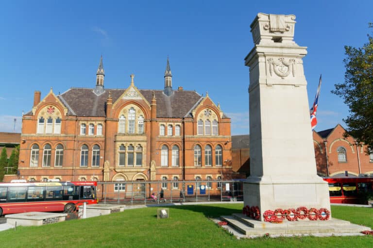 A historic red brick building with a grand facade and towers stands behind a white stone war memorial adorned with wreaths. Nearby, a Hair Loss Clinic offers discreet services. A Union Jack flag flutters beside red buses parked on the side. The sky is clear and blue.