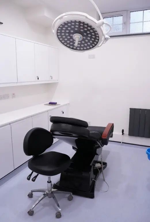 A modern medical examination room designed for hair transplants in Manchester, featuring a reclining black exam chair, an adjustable stool, white cabinets, an overhead surgical light, and a large window letting in natural light.